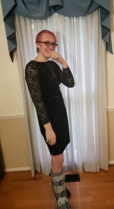 A young white person with pink hair, a black velvet dress, and a cam boot on one foot laughing.